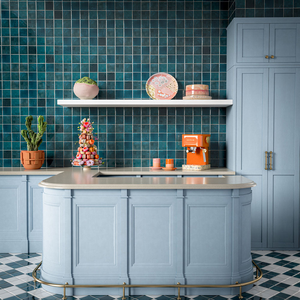 Kitchen splashback featuring Levent Blue Square 10cm x 10cm gloss porcelain wall tiles, showcasing rich blue tones with subtle handmade-effect shade variation, paired with pale blue cabinetry, light stone worktops, open shelving and vibrant contemporary accessories.