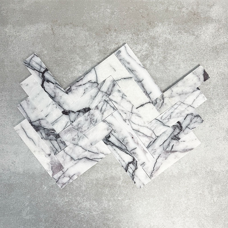 Arrangement of Fayrouz Lilac 5x20cm natural marble tiles with a honed finish, displayed in a herringbone layout, showcasing a soft white base with flowing lilac and grey veining, highlighting natural variation and adding movement and elegance to the design.