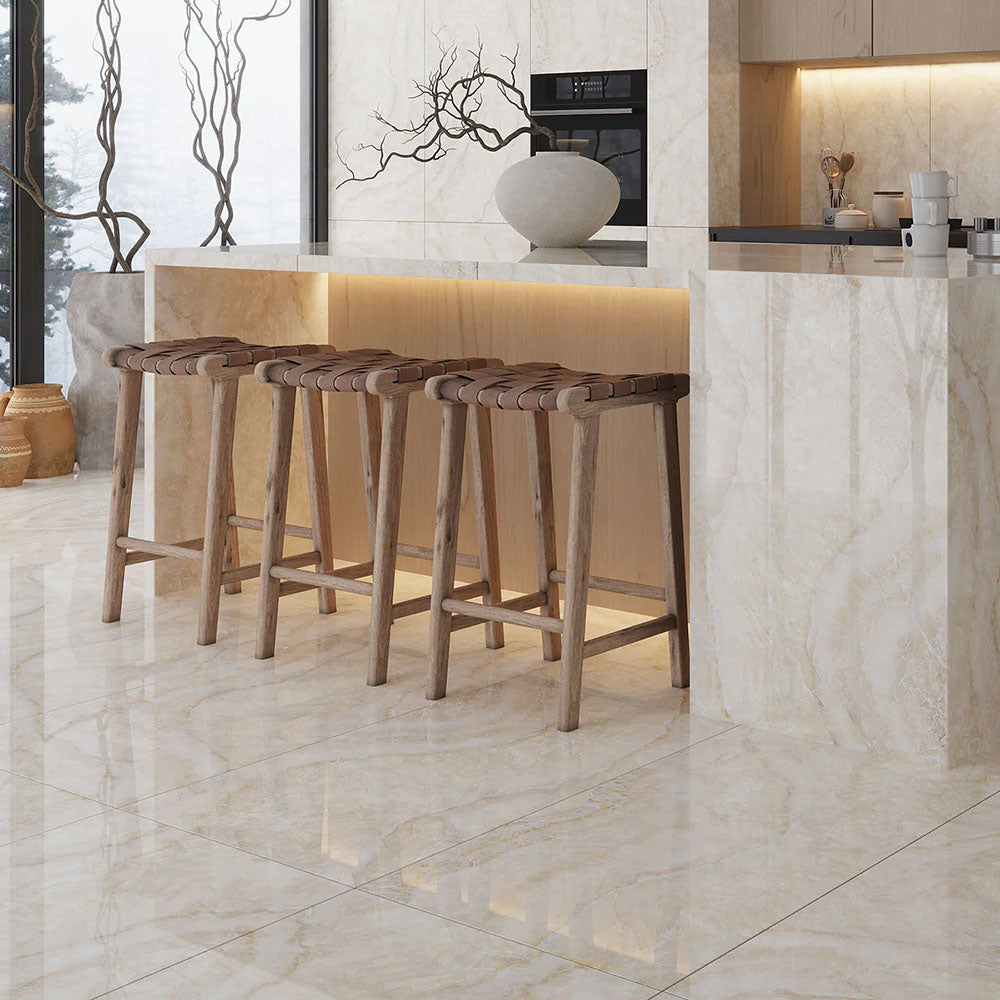 Open-plan kitchen interior featuring Lumen Precious Stone Ivory 60 × 120cm polished porcelain wall and floor tiles, showcasing warm ivory and cream gemstone-inspired tones with crystalline layering, delicate natural veining, and a reflective high-gloss finish, paired with light wood cabinetry, a statement island, woven leather bar stools, and soft ambient lighting.