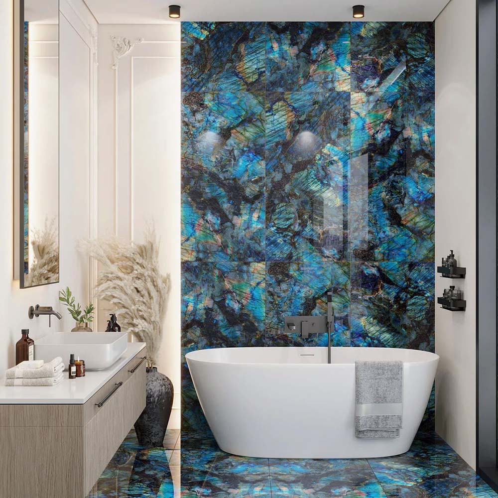 Bathroom interior featuring Lumen Precious Stone Labradorite 60 × 120cm polished porcelain wall and floor tiles, showcasing vivid blue, teal, and charcoal gemstone-inspired detailing with an iridescent high-gloss finish, paired with a freestanding white bath and brushed metal fittings for a dramatic luxury look.
