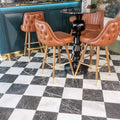 Interior setting featuring Fayrouz White 15.2x15.2cm natural marble tiles with a honed finish, installed in a classic chequerboard floor with black marble, styled with tan leather bar stools and a dark statement table.