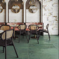 Contemporary restaurant interior featuring Mediterraneo Green 2cm outdoor slabs 50 × 100cm, showcasing deep green stone-effect porcelain tiles, paired with cane dining chairs, brass lighting, and floral marble-effect feature walls.
