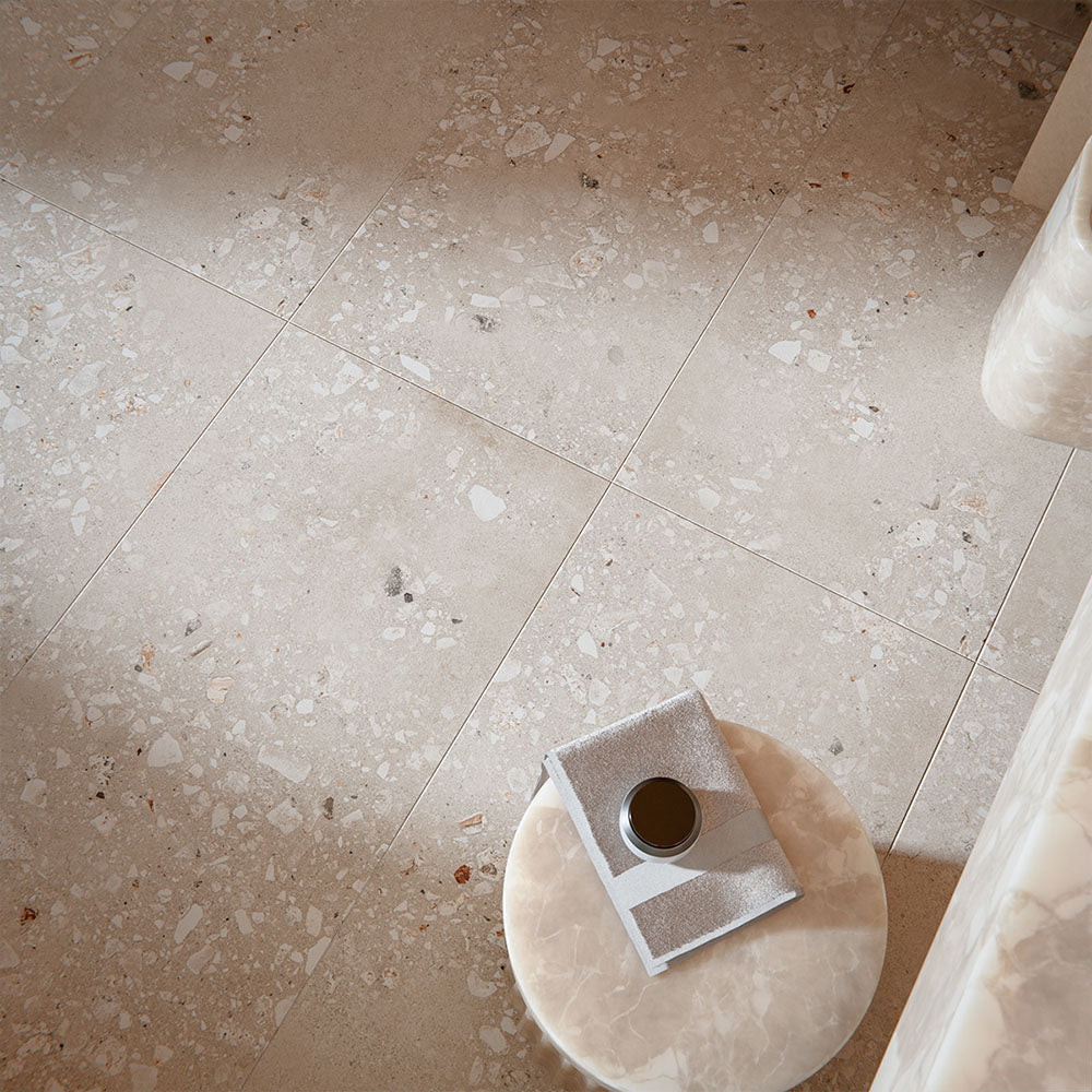 Contemporary interior featuring Melange Sand Cosmo Stone wall and floor tiles 60 × 120cm, showcasing warm sand-toned stone-effect detailing with terrazzo-inspired aggregate texture, paired with soft neutral furnishings and marble accents.
