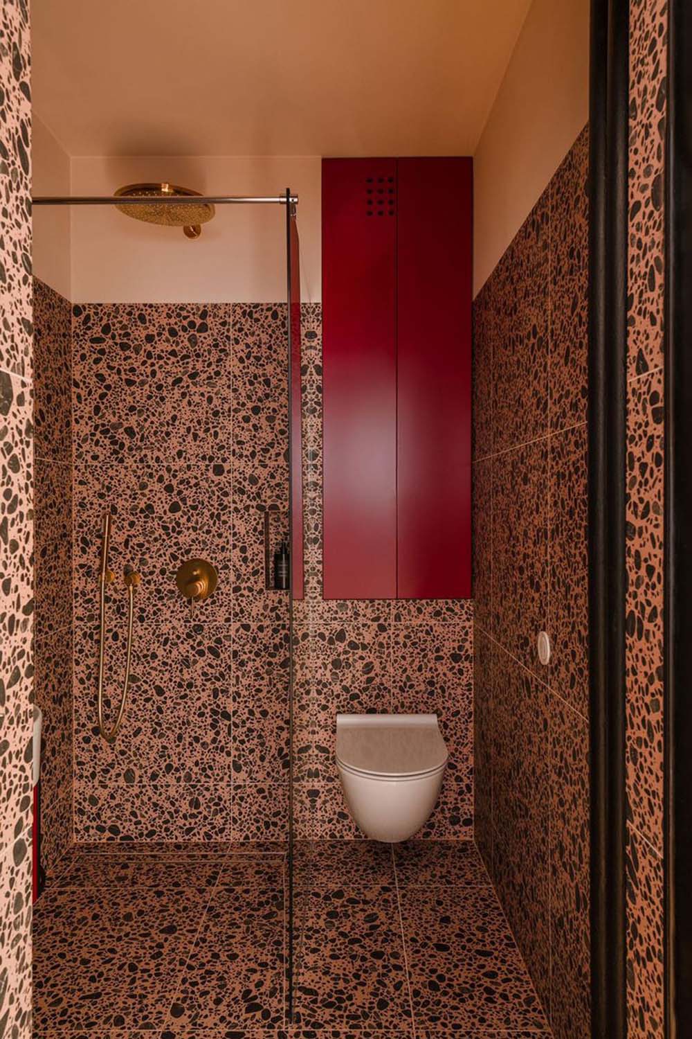 Bold bathroom interior featuring Melody POP Terrazzo Pink 60 × 60cm porcelain tiles, showcasing a striking terrazzo-effect design with oversized dark stone fragments set against warm pink tones, paired with black fittings and vibrant red vanity units for a playful contemporary look.