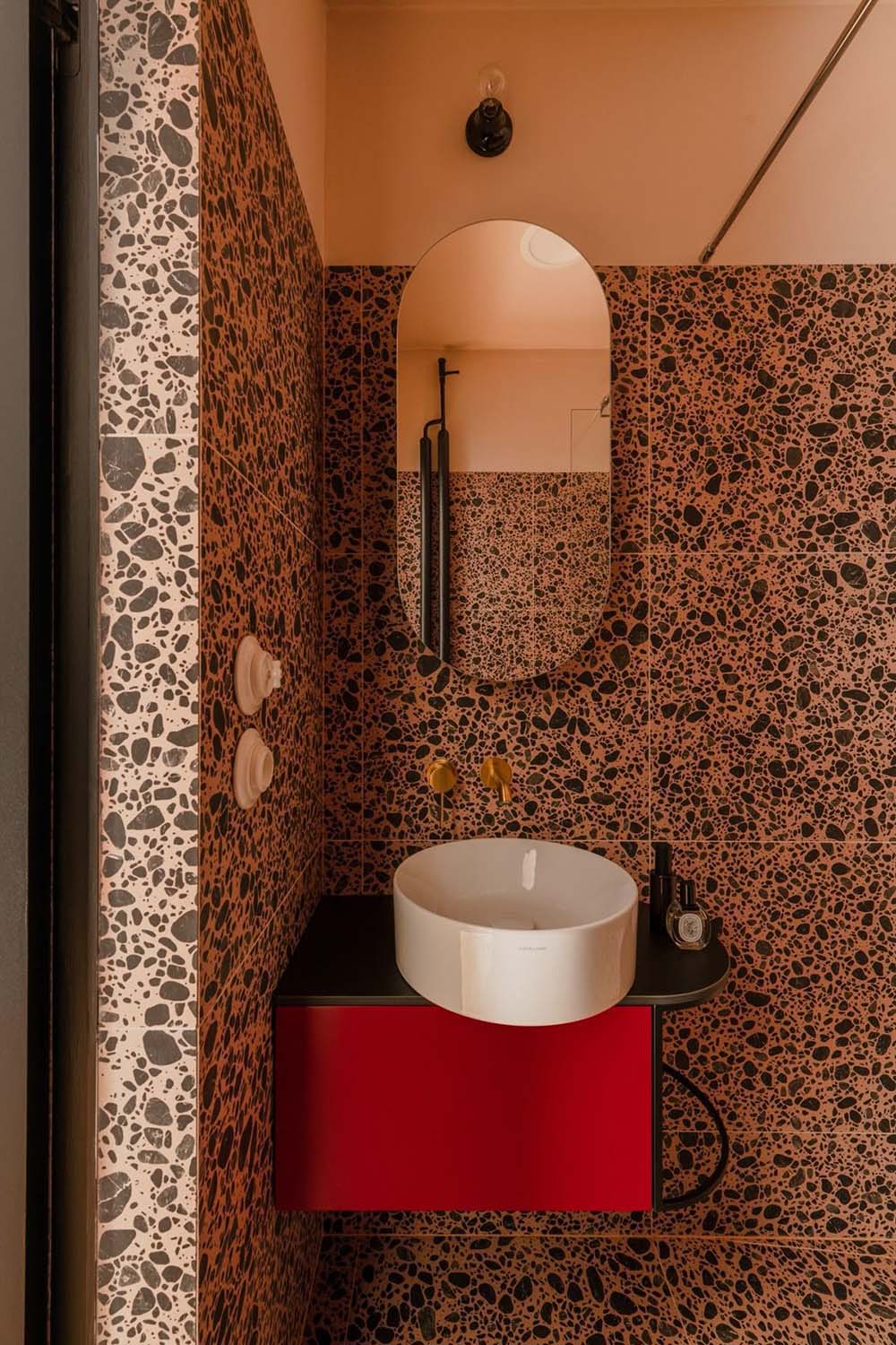Bold bathroom interior featuring Melody POP Terrazzo Pink 60 × 60cm porcelain tiles, showcasing a striking terrazzo-effect design with oversized dark stone fragments set against warm pink tones, paired with black fittings and vibrant red vanity units for a playful contemporary look.