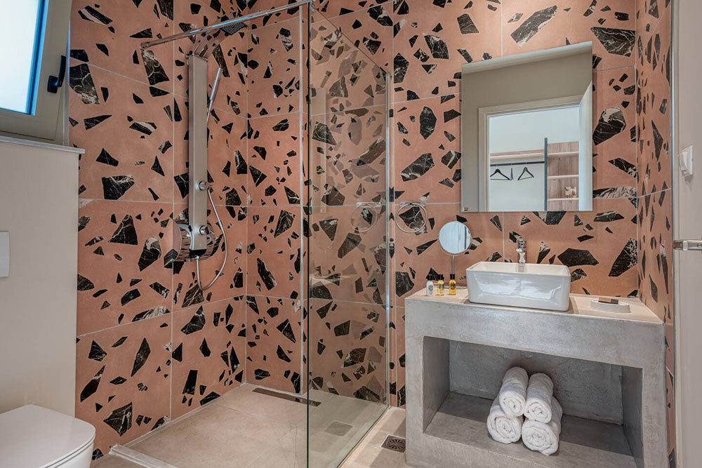 Modern bathroom featuring Melody ROCK Terrazzo Pink 60 × 60cm porcelain wall tiles, showcasing oversized black marble-effect fragments across a warm pink terrazzo-inspired surface, paired with concrete styling, minimalist fittings, and crisp white sanitaryware for a bold contemporary finish.