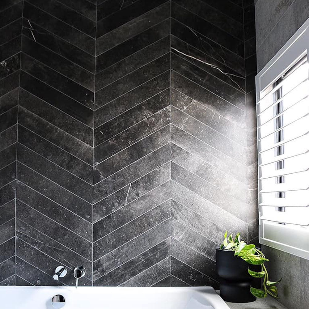 Lifestyle image of Monzo Stone Chevron Nite 10x53cm dark chevron porcelain wall tiles in deep charcoal tones, featuring subtle stone-effect texture and delicate veining, styled within a contemporary bathroom with dramatic contrast and modern black accents.