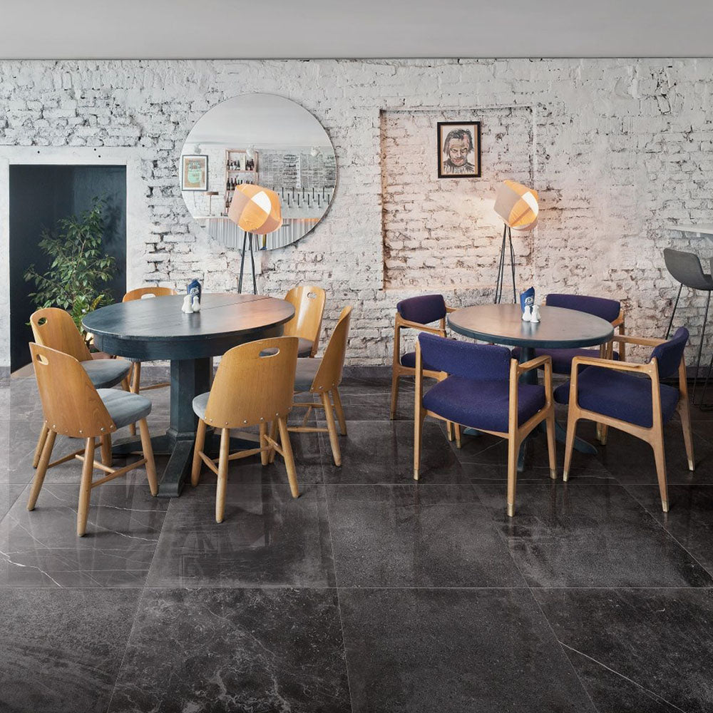 Lifestyle image of Monzo Stone Nite 60x60cm dark stone-effect porcelain floor tiles in deep charcoal tones, featuring subtle veining and varied natural texture, styled within a contemporary café interior with industrial-inspired décor and warm wood furnishings.