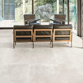 Living room featuring Monzo Stone Silk 60x60cm stone-effect porcelain floor tiles in soft ivory tones, styled with black leather lounge chairs, warm wood furniture, and floor-to-ceiling glazing for a modern interior look.
