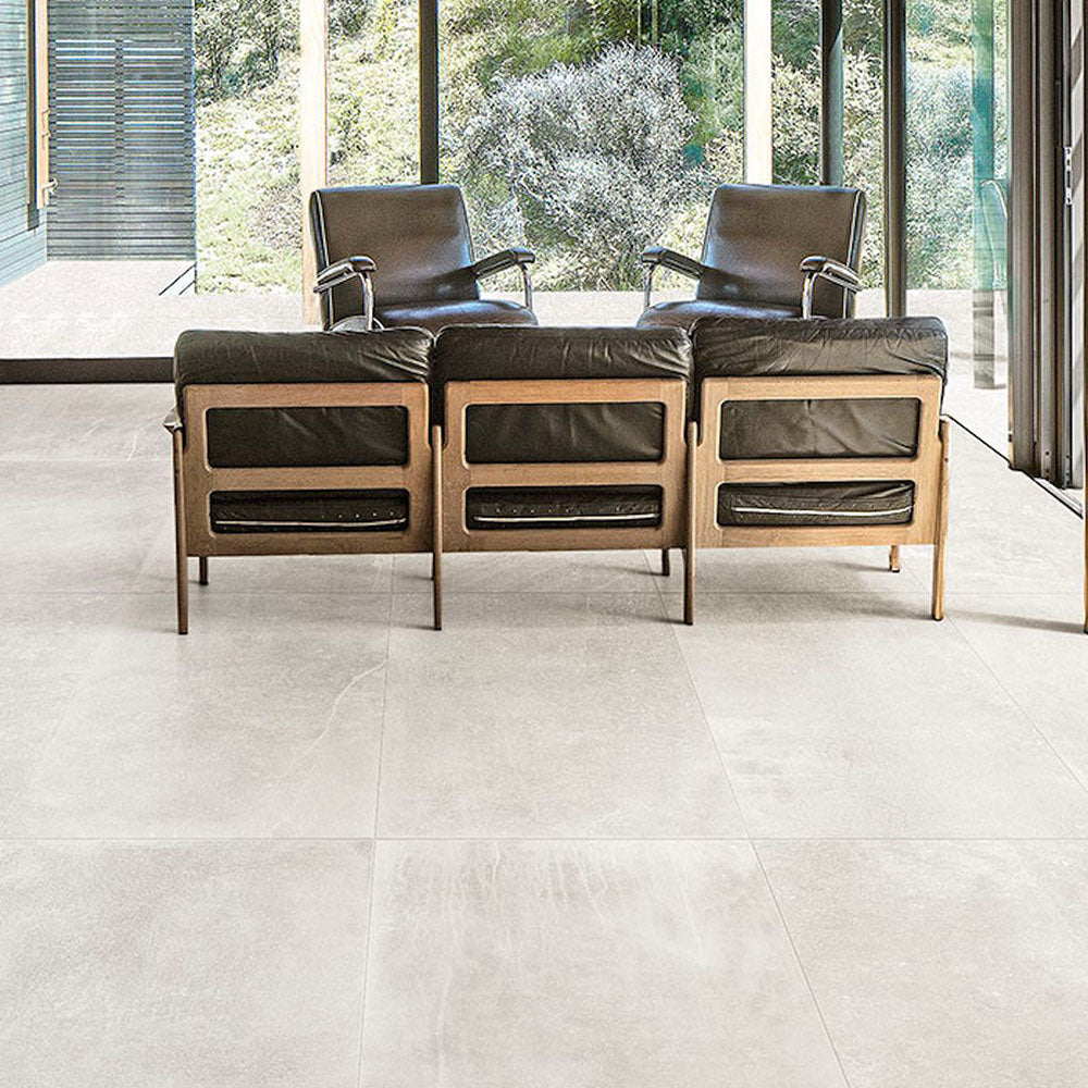 Living room featuring Monzo Stone Silk 60x60cm stone-effect porcelain floor tiles in soft ivory tones, styled with black leather lounge chairs, warm wood furniture, and floor-to-ceiling glazing for a modern interior look.