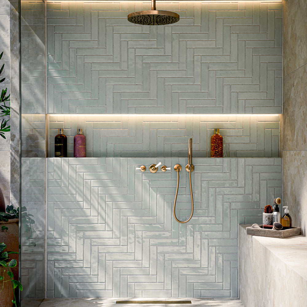 Bathroom featuring Nomad Mint Green Brick 6x24.6cm industrial-style wall tiles, laid in a herringbone pattern and showcasing a glossy mint green finish, paired with brass shower fittings, recessed shelving and warm ambient lighting.