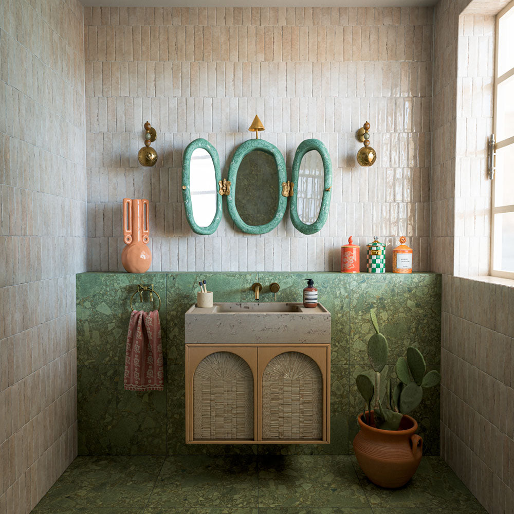 Bathroom featuring Nomad Oatmeal industrial style wall tiles, showcasing soft oatmeal and beige tones with subtle surface variation and a glossy finish. Installed in a vertical stacked layout, the tiles create texture and depth while complementing the rich green stone-effect surfaces, natural materials and brass accents. The result is a warm, characterful space that blends contemporary design with earthy Mediterranean-inspired styling.