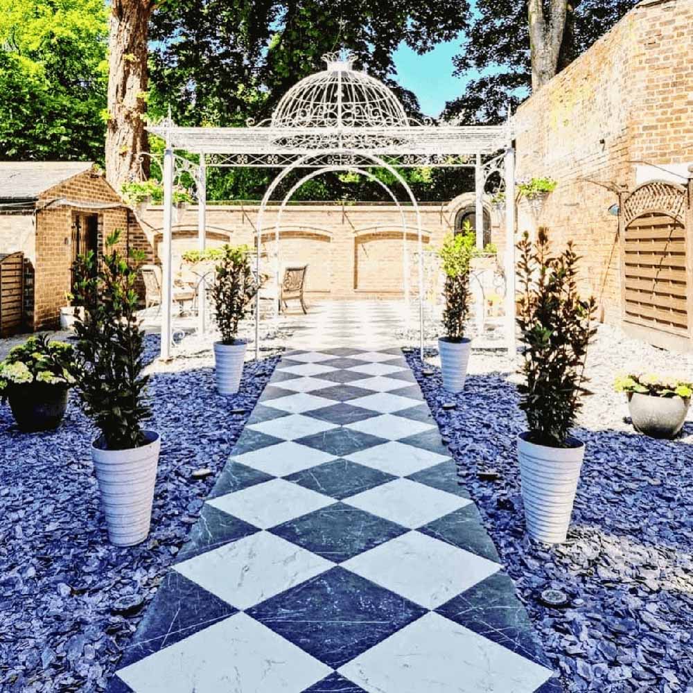 Outdoor garden walkway featuring Florence Grand Marquina Black 50x50cm marble-effect porcelain floor tiles, laid in a dramatic black and white diamond checkerboard pattern and styled with potted greenery, gravel borders and decorative metal archways for an elegant outdoor statement.
