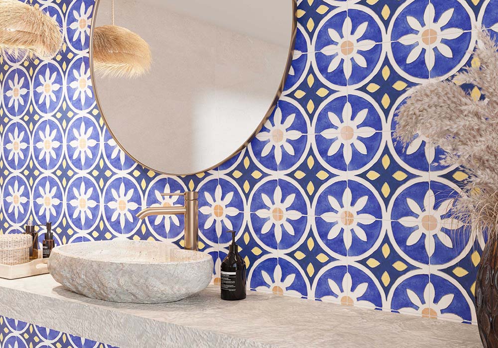 Bathroom featuring Peniscola Blue and Yellow patterned wall and floor tiles, showcasing a bold Mediterranean-inspired floral geometric design in vibrant cobalt blue, warm yellow and crisp white tones, paired with a floating stone vanity, round mirror, woven pendant lights, natural baskets and soft neutral finishes for a bright, characterful interior.