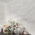 Living room featuring Petra Avorio Ivory stone marble effect porcelain wall tiles in a 60cm x 120cm format, showcasing soft ivory, pearl white and light grey veining inspired by natural stone, paired with colourful glass vases, fresh floral arrangements and contemporary styling to create a bright, elegant interior.