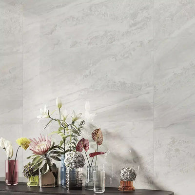 Living room featuring Petra Avorio Ivory stone marble effect porcelain wall tiles in a 60cm x 120cm format, showcasing soft ivory, pearl white and light grey veining inspired by natural stone, paired with colourful glass vases, fresh floral arrangements and contemporary styling to create a bright, elegant interior.