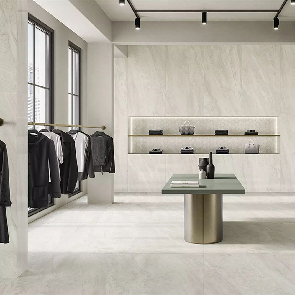 Retail showroom featuring Petra Beige stone marble effect porcelain wall and floor tiles in a 30cm x 60cm format, showcasing warm beige, ivory and soft cream veining inspired by natural stone, paired with minimalist shelving, contemporary display furniture, black metal clothing rails and a bright, modern commercial interior.