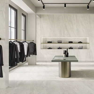 Retail showroom featuring Petra Beige stone marble effect porcelain wall and floor tiles in a 30cm x 60cm format, showcasing warm beige, ivory and soft cream veining inspired by natural stone, paired with minimalist shelving, contemporary display furniture, black metal clothing rails and a bright, modern commercial interior.