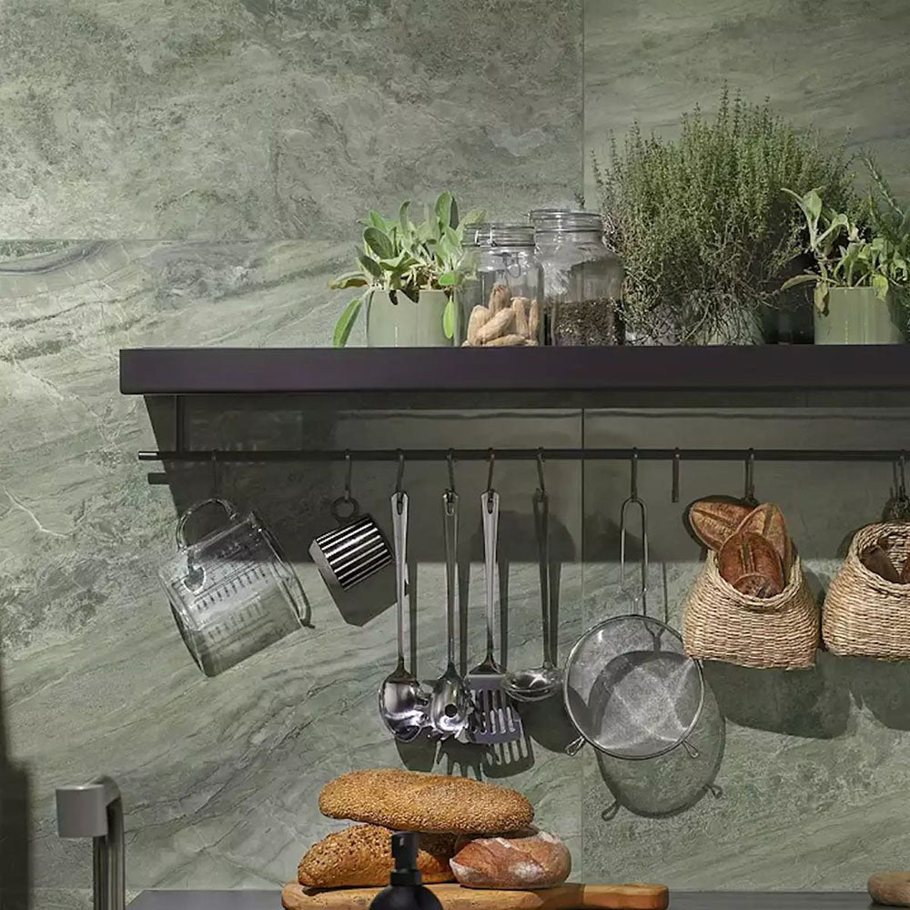 Kitchen featuring Petra Verde Green stone marble effect porcelain wall tiles in a 30cm x 60cm format, showcasing flowing green, sage and soft grey veining inspired by natural stone, paired with dark cabinetry, black shelving, hanging kitchen utensils, natural greenery and woven baskets for a contemporary organic interior.