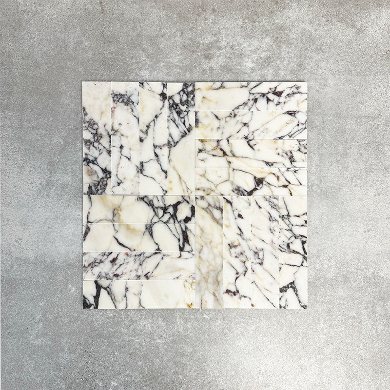 Arrangement featuring Fayrouz Picasso 5x20cm natural marble tiles with a honed finish, laid in a basketweave pattern to create visual texture and structure, showcasing a white base with bold grey, green and warm gold veining and natural variation across each piece.