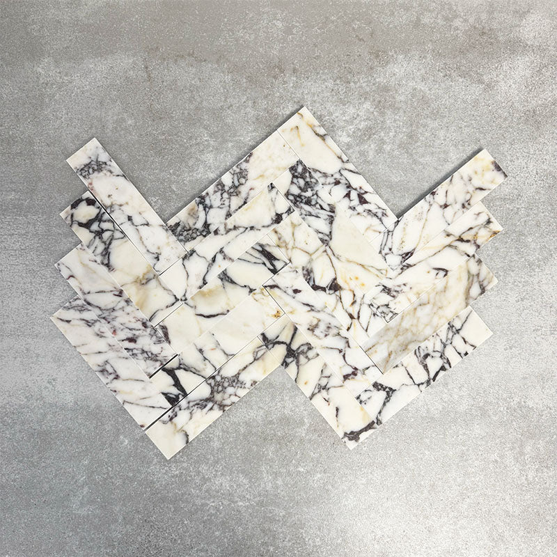 Arrangement featuring Fayrouz Picasso 5x20cm natural marble tiles with a honed finish, laid in a herringbone pattern to create directional movement, showcasing a white base with bold grey, green and warm gold veining and natural variation across each piece.