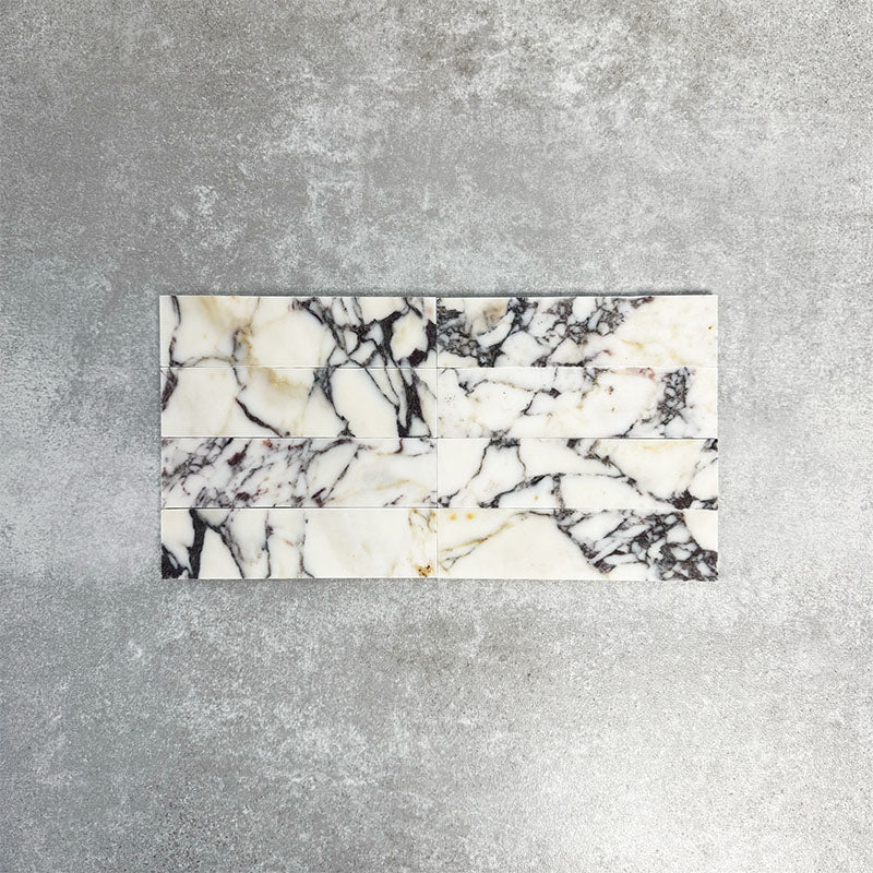 Arrangement featuring Fayrouz Picasso 5x20cm natural marble tiles with a honed finish, laid in a horizontal brick pattern to create a clean, linear look, showcasing a white base with bold grey, green and warm gold veining and natural variation across each piece.