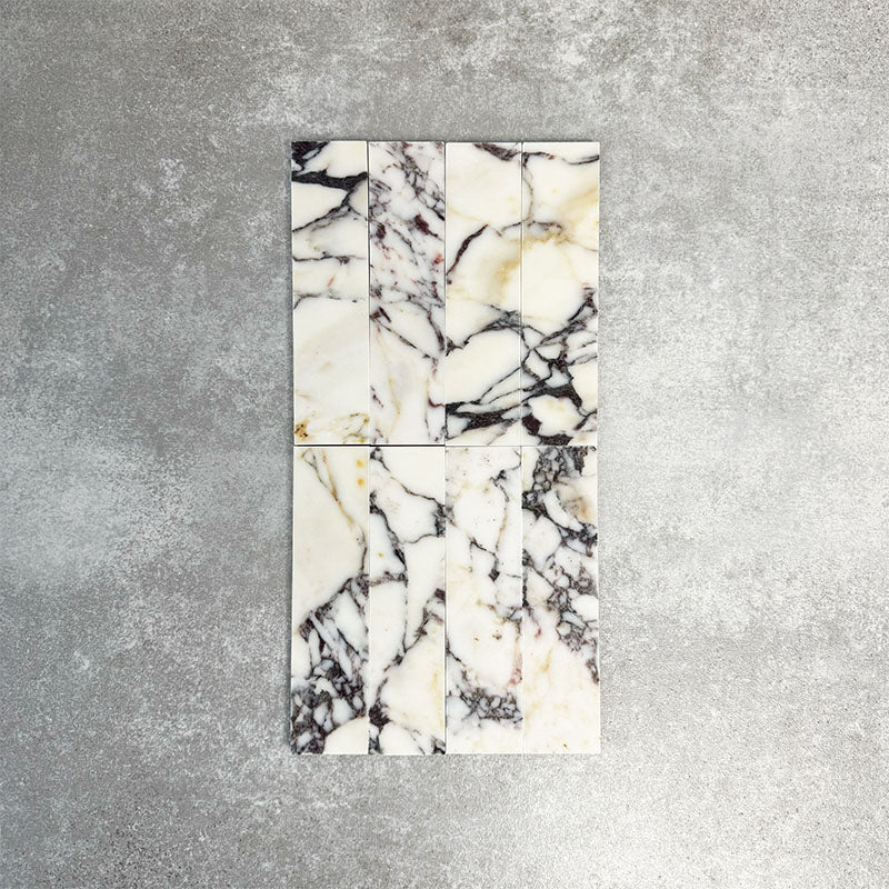 Arrangement featuring Fayrouz Picasso 5x20cm natural marble tiles with a honed finish, displayed in a vertical layout to emphasise a white base with bold grey, green and warm gold veining, showcasing natural variation and flowing movement across each piece.