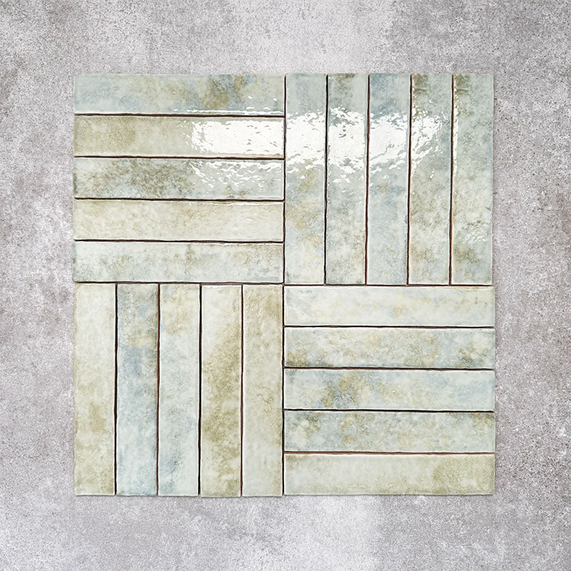 Product display of Pottery Brick Agave Green 5x25cm glossy wall tiles in soft green and sage tones, arranged in a basketweave layout with handcrafted edges and subtle tonal variation against a textured concrete-style backdrop.