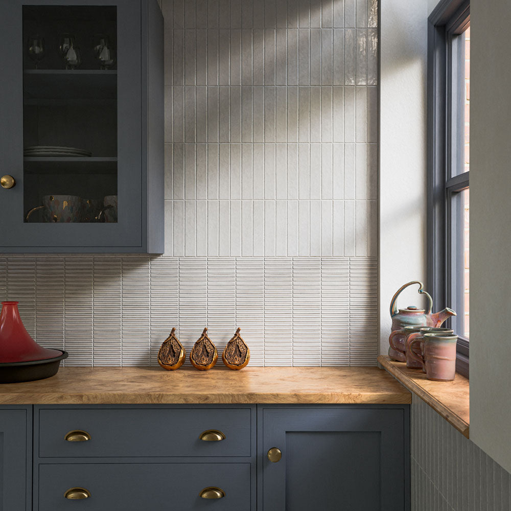 Kitchen splashback featuring Pottery Decor Blanco 12.5 × 25cm ceramic wall tiles, combining textured linear relief tiles with glossy slim brick tiles in soft white tones, paired with deep blue cabinetry, warm wood worktops, and brass accents for a layered contemporary design.