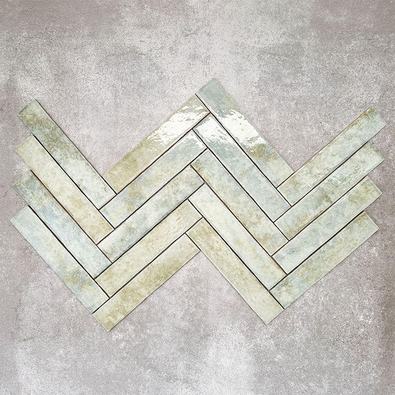 Product display of Pottery Brick Agave Green 5x25cm glossy wall tiles in soft green and sage tones, arranged in a herringbone layout with handcrafted edges and subtle tonal variation against a textured concrete-style backdrop.