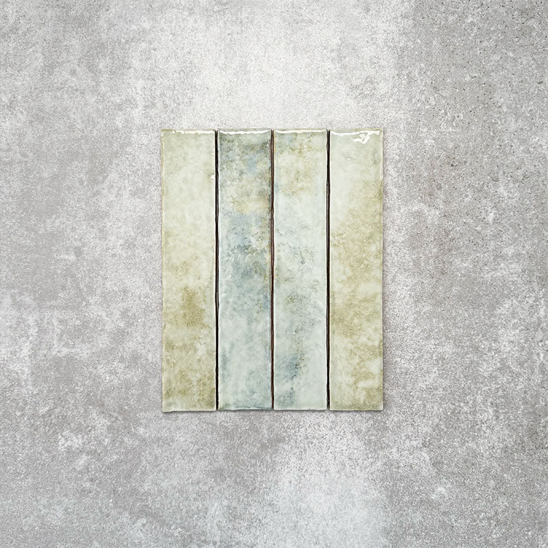 Product display of Pottery Brick Agave Green 5x25cm glossy wall tiles in soft sage and green tones, featuring handcrafted edges and subtle tonal variation against a textured concrete-style backdrop.