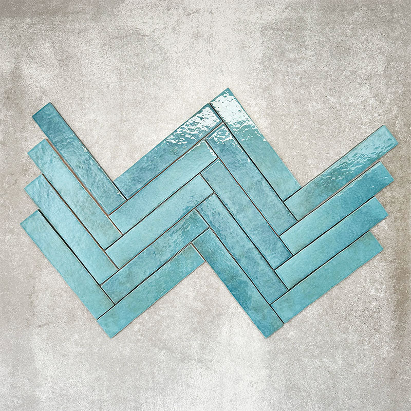 Product display of Pottery Brick Azul Blue 5x25cm glossy wall tiles in vibrant blue tones, arranged in a herringbone layout with handcrafted edges and tonal variation against a textured concrete-style backdrop.