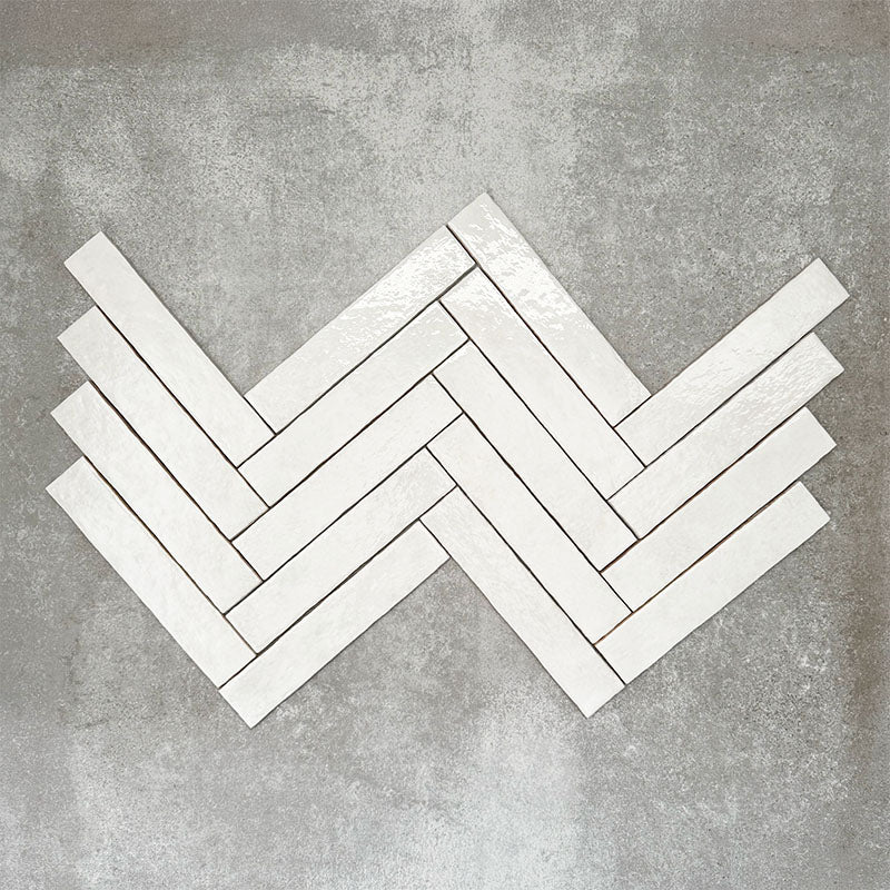 Product display of Pottery Brick Blanco 5x25cm glossy wall tiles in soft white tones, arranged in a herringbone layout with handcrafted edges and subtle surface variation against a textured concrete-style backdrop.