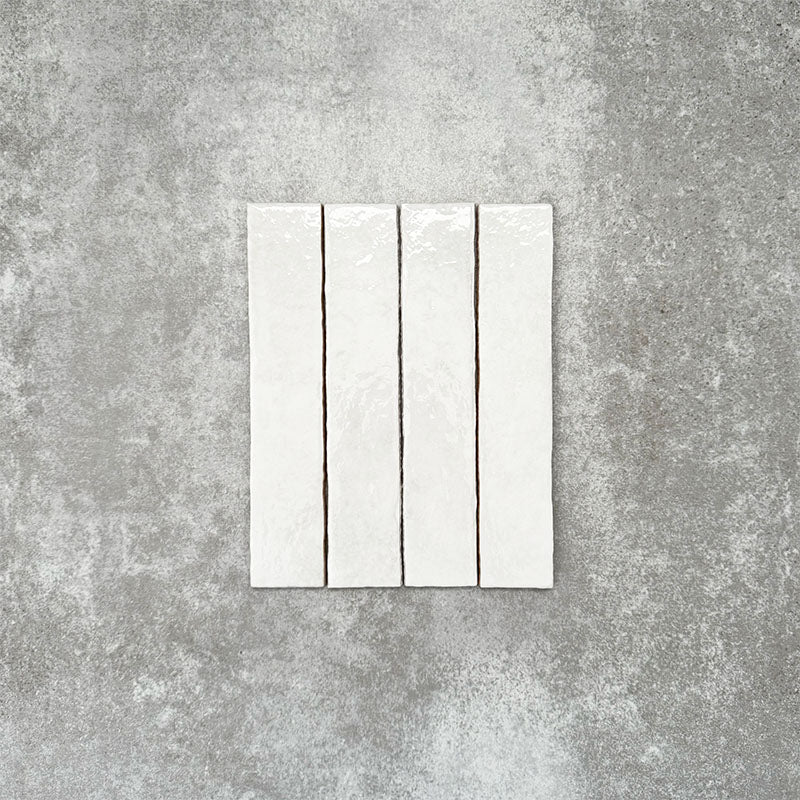 Product display of Pottery Brick Blanco 5x25cm glossy wall tiles in soft white tones, featuring handcrafted edges and subtle surface variation against a textured concrete-style backdrop.