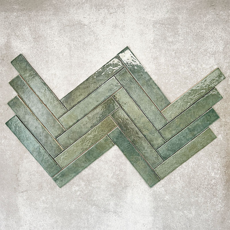 Product display of Pottery Brick Forest Green 5x25cm glossy wall tiles in deep green tones, arranged in a herringbone layout with handcrafted edges and rich tonal variation against a textured concrete-style backdrop.
