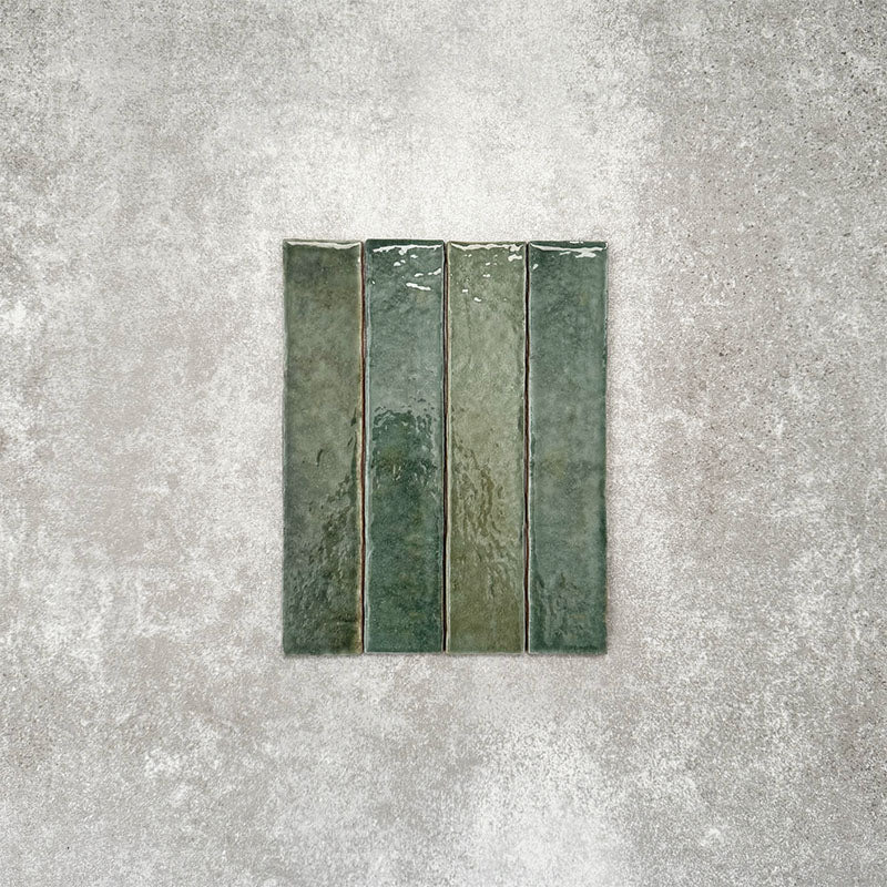 Product display of Pottery Brick Forest Green 5x25cm glossy wall tiles in rich forest green tones, featuring handcrafted edges and tonal variation against a textured concrete-style backdrop.