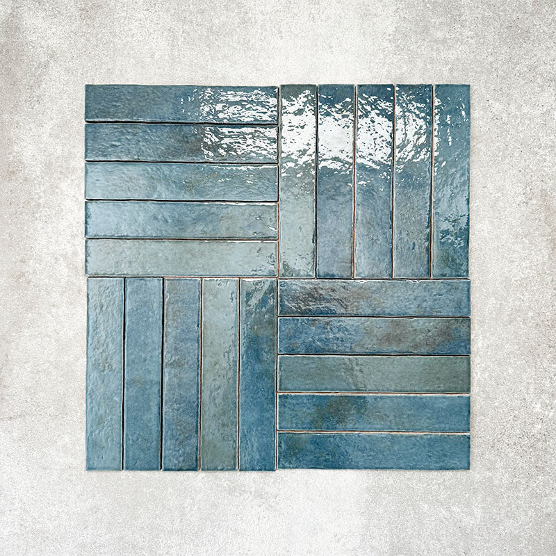 Product display of Pottery Brick Prussian 5x25cm glossy wall tiles in deep blue tones, arranged in a basketweave layout with handcrafted edges and rich tonal variation against a textured concrete-style backdrop.