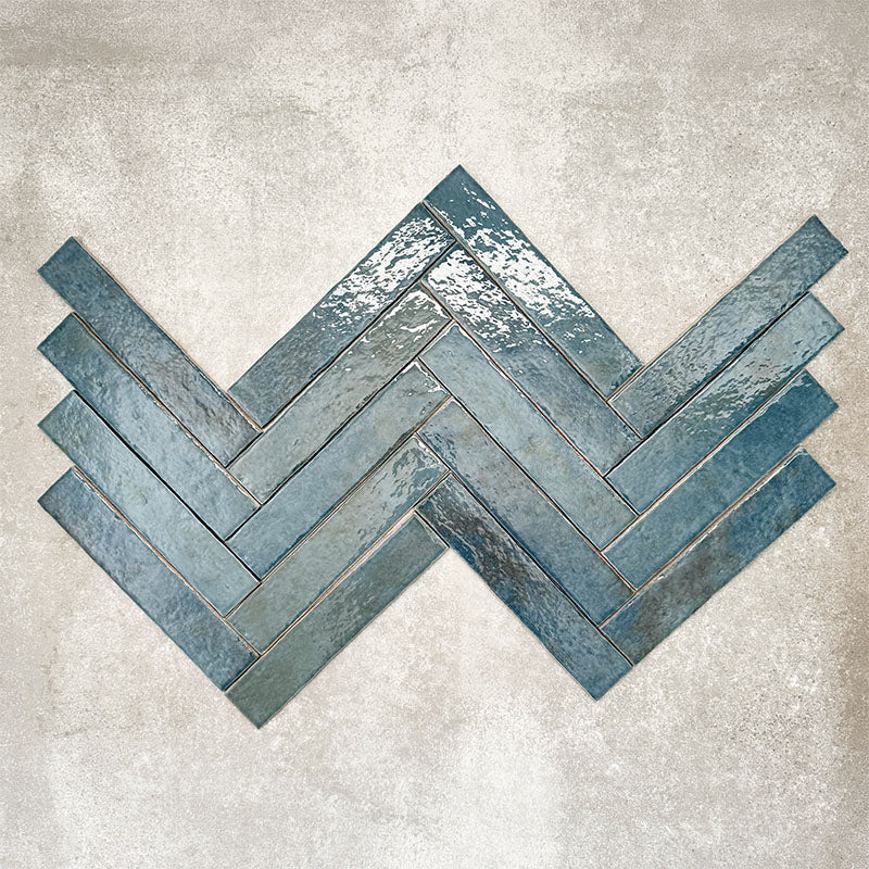 Product display of Pottery Brick Prussian 5x25cm herringbone wall tiles in deep blue tones, arranged in a basketweave layout with handcrafted edges and rich tonal variation against a textured concrete-style backdrop.