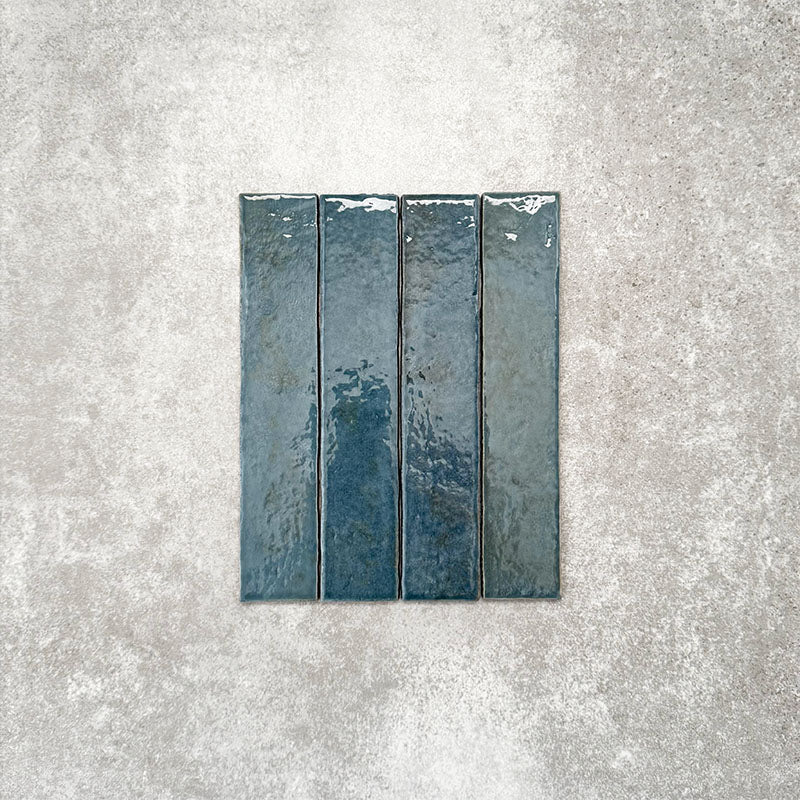 Product display of Pottery Brick Prussian 5x25cm glossy wall tiles in deep Prussian blue tones, featuring handcrafted edges and rich tonal variation against a textured concrete-style backdrop.