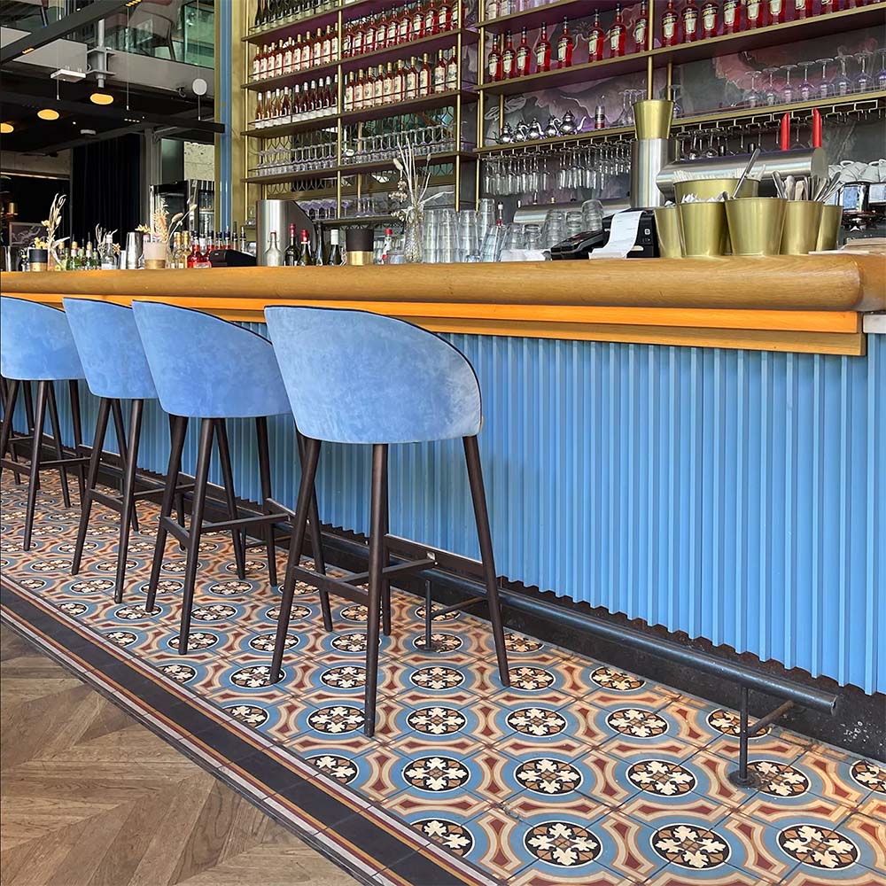 Restaurant seating area featuring London Streets Carnegie Hall 20x20cm Victorian Moroccan style floor tiles, styled with pink upholstered chairs, wooden tables, navy curtains and large windows creating a colourful patterned café interior.