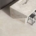 Contemporary kitchen featuring Riad Avorio Neutra Stone 30x60cm stone-effect wall and floor tiles, showcasing warm ivory and beige tones, paired with a matching waterfall island, black bar stools and minimalist finishes.