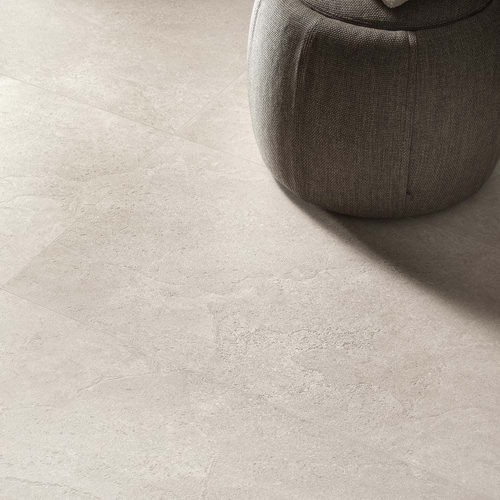 Close-up of Riad Avorio Neutra Stone 30x60cm stone-effect floor tiles, showcasing warm ivory and beige tones with delicate natural stone-inspired veining, paired with a soft upholstered pouffe in a contemporary interior.