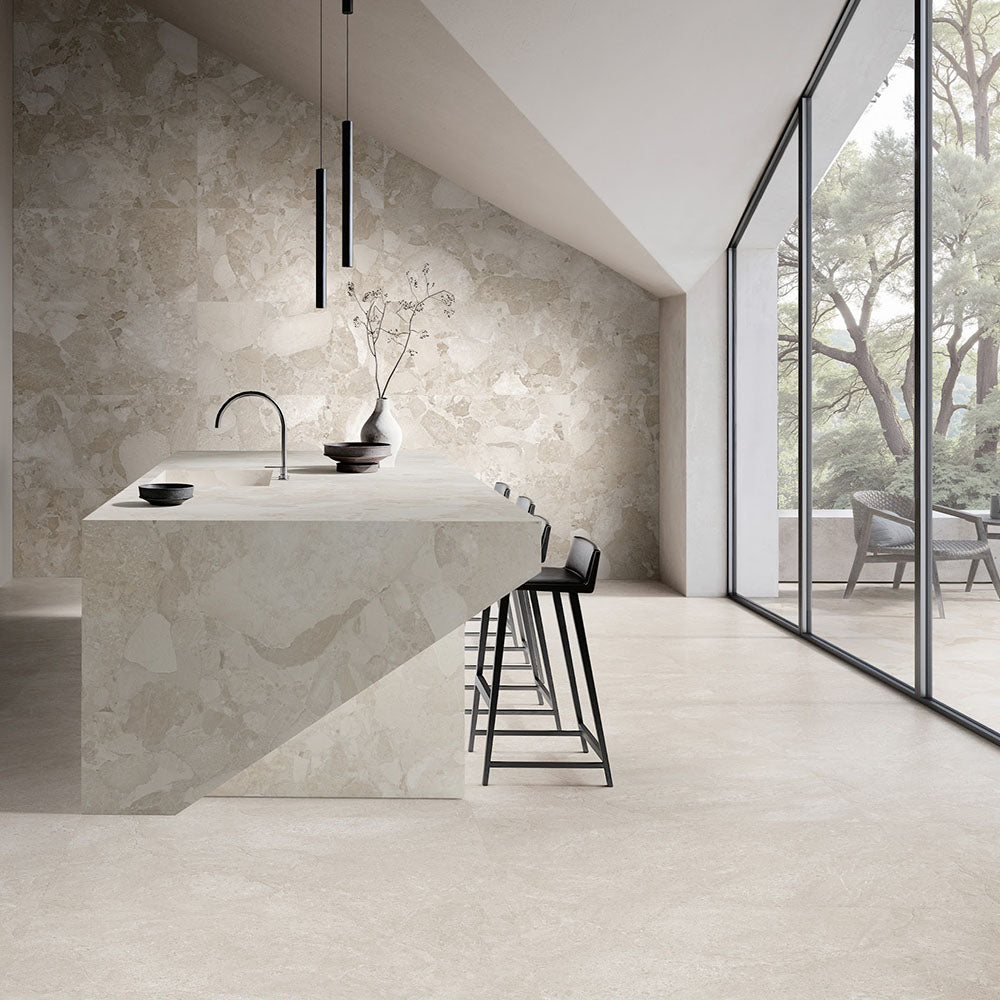 Contemporary kitchen featuring Riad Avorio Sassi Stone 30x60cm stone-effect wall and floor tiles, showcasing warm ivory and beige tones with natural stone-inspired detailing, paired with a matching waterfall island, black bar stools and floor-to-ceiling glazing.