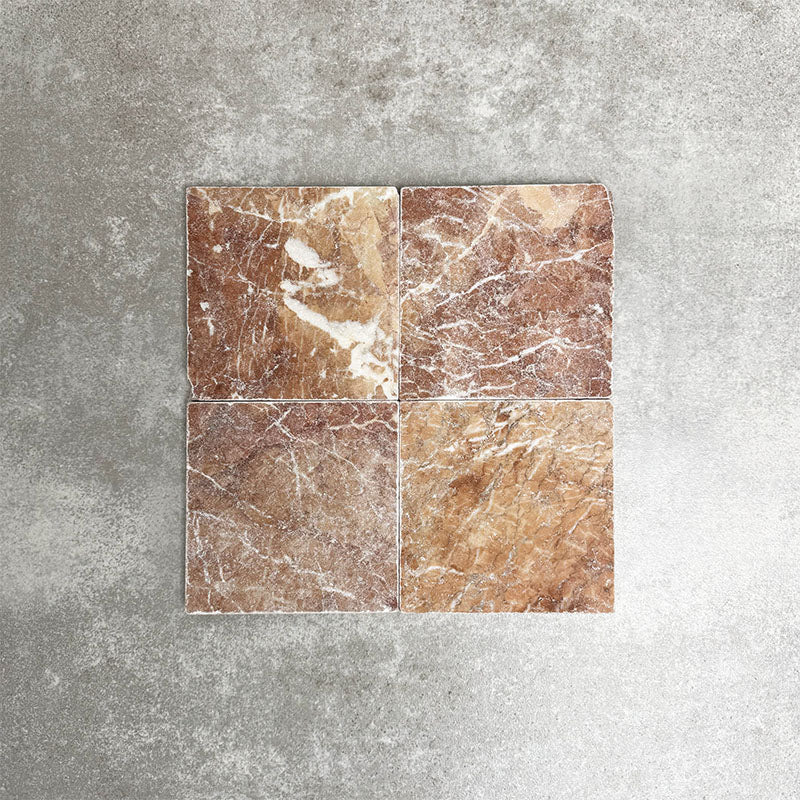 Four Fayrouz Rosso 15.2x15.2cm natural marble tiles with a tumbled finish arranged together, showcasing warm red tones, distinctive veining and natural variation against a neutral textured background.