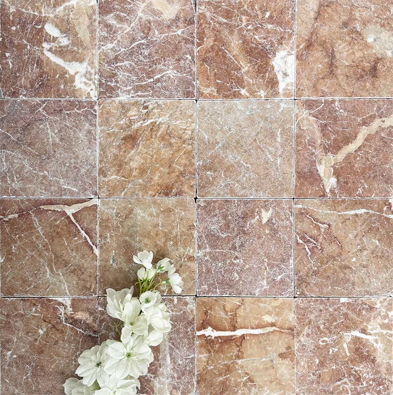 Flat lay of Fayrouz Rosso 15.2x15.2cm natural marble tiles with a tumbled finish, featuring warm red tones, natural veining and surface variation, styled with soft white flowers for contrast.