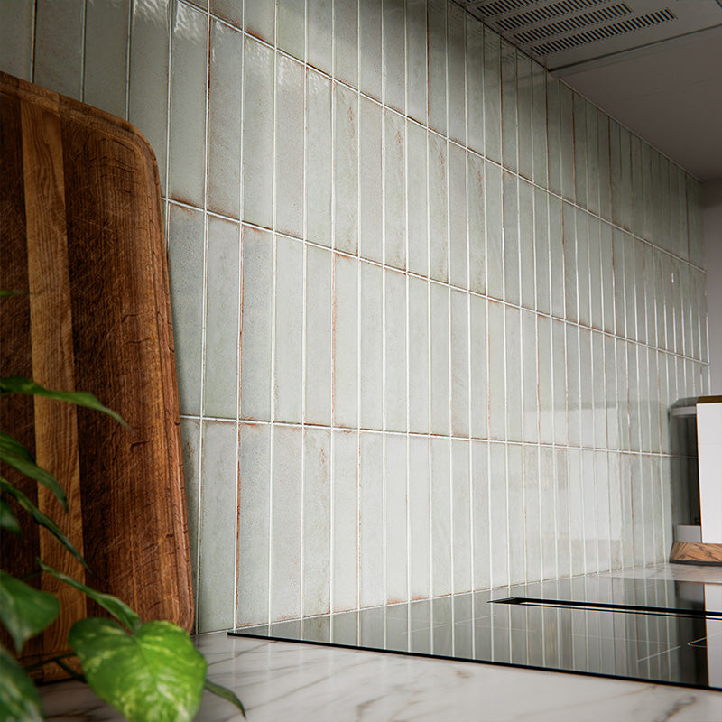Puzzle Breeze Green Brick Rectified 5x20cm porcelain wall and floor tiles styled across a curved kitchen island and splashback, showcasing soft breeze green tones with subtle speckling and gentle shade variation. The elongated brick format enhances the island’s rounded form, while pairing beautifully with warm wood cabinetry, marble worktops and brushed brass fixtures for a calm, contemporary finish.