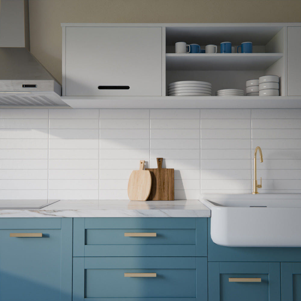 Modern kitchen featuring Bora Bora White Sand Brick ceramic wall tiles in a 6.5 × 32.8cm slim brick format with a flux glaze finish, laid horizontally as a splashback behind blue cabinetry, paired with white worktops, open shelving, and brass tapware for a clean, contemporary look.
