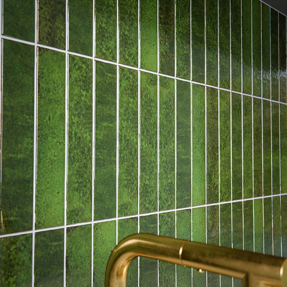 Close-up of Bora Bora Tourmaline Green Brick wall tiles in 6.5x32.8cm format with a flux glaze finish, installed vertically to showcase rich green tonal variation, glossy texture and organic surface movement beside brass fittings.