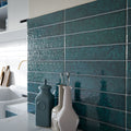 Kitchen splashback featuring Bora Bora Amun Blue Brick ceramic wall tiles in a 6.5 × 32.8cm slim brick format with a flux glaze finish, laid horizontally to showcase rich blue-green tonal variation, paired with teal cabinetry, marble-effect worktops, and brass fixtures for a bold, contemporary look.