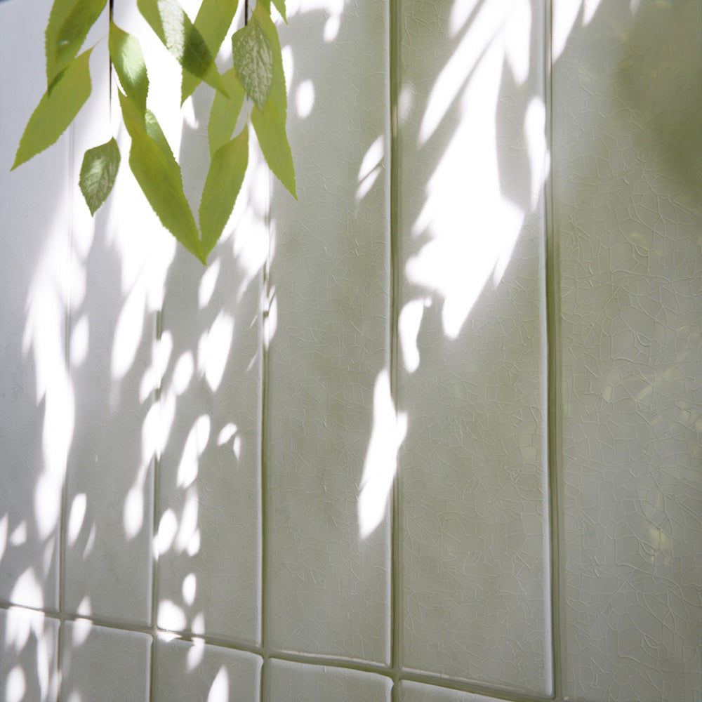 Modern bathroom featuring Bora Bora White Sand Brick ceramic wall tiles in a 6.5 × 32.8cm slim brick format with a flux glaze finish, laid vertically and horizontally to create a soft, textured feature wall, paired with a freestanding bath, brass tapware, natural light, and greenery for a calm, spa-like atmosphere.